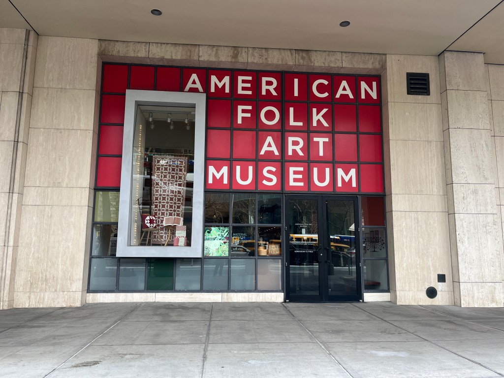 Front entrance of the American Folk Art Musuem, with large red sign with white lettering.