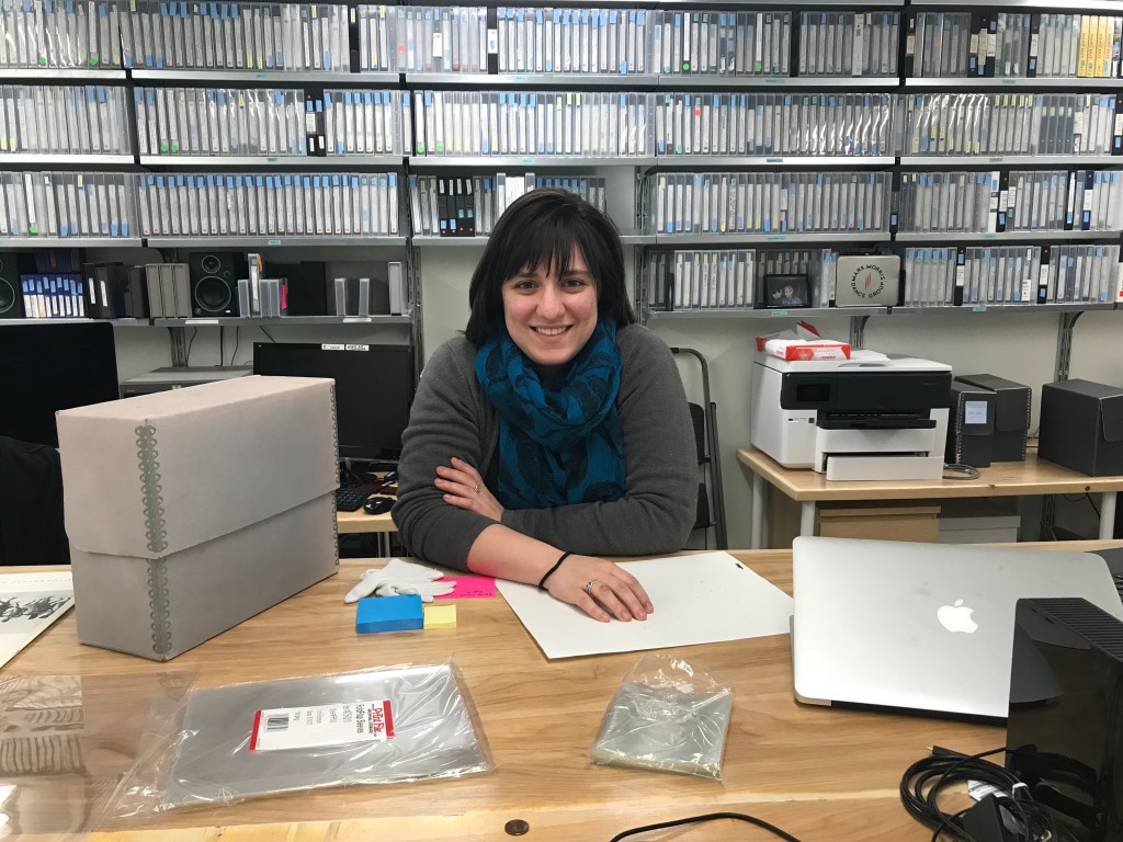 Regina Carra sitting at a table, wearing a grey cardigan sweater and blue scarf with feather pattern. On the table are archive supplies--plastic sleeves, white cotton gloves, blue post-it notes, a grey box, and silver computer. In the background are shelves of VHS tapes.