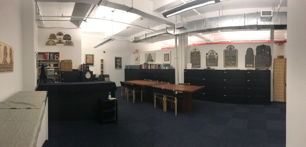 A large archive room with dark blue carpet. Two wooden tables in the middle of the room in front of black file cabinets. Posters and gravestone rubbings are hung on the walls around the room.