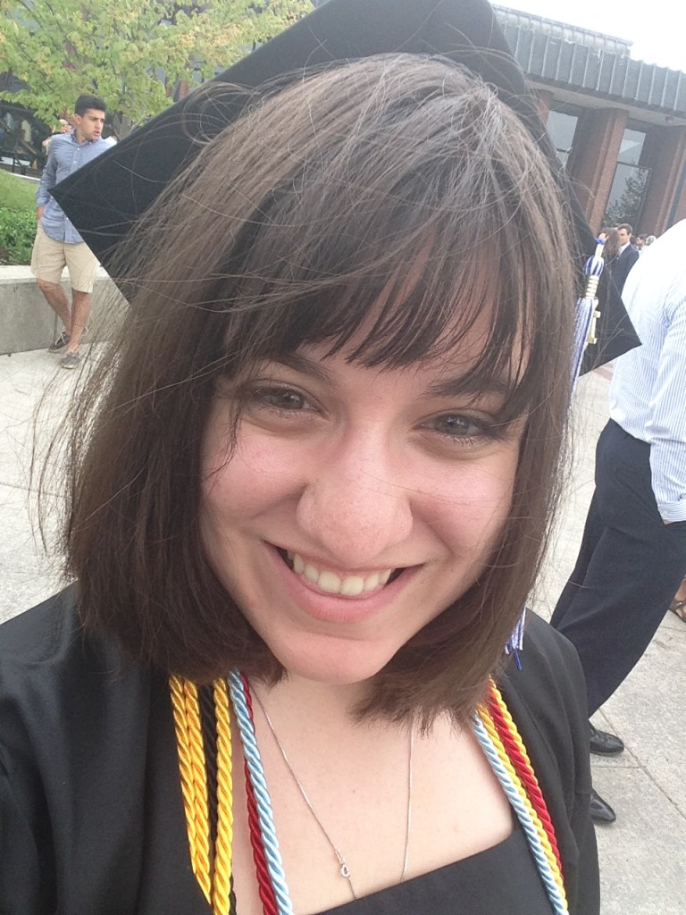 A selfie of Regina Carra smiling at college graduation. She is wearing a black cap and grown with gold, red, and blue tassels around her neck. In this photo she is a 22 year old white woman, with chin-length brown hair.