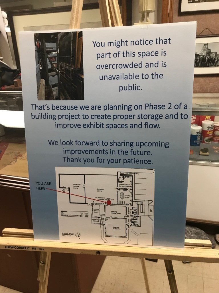 A sign on a wooden easel. The sign is blue with blue lettering. It includes a photo of a cramped storage room and a blue print of new museum rooms to be built. The sign says: "You might notice that part of this space is overcrowed and is unavailable to the public. That's because we are planning on Phase 2 of a building project to create proper storage and to improve exhibit spaces and flow. We look forward to sharing upcoming improvements in the future. Thank you for your patience."