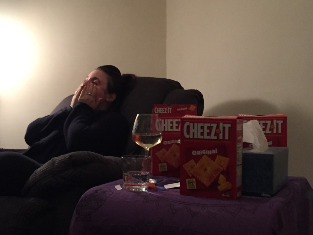 Photo of Regina Carra sitting in a green recliner chair rubbing her eyes with her hands in exhaustion. To the right of the chair is a wide-table with purple table cloth. On the table is a glass of water, glass of white wine, three red Cheez-It boxes, and a tissue box.