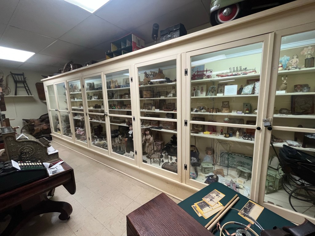 A large white cabinet with glass doors. Little toys and figurines inside the case are on display, such as dolls, toy ships, and games.