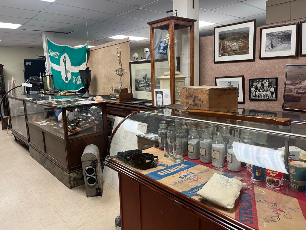 Artifacts everywhere in the background. Wooden display case with jars and bags of salt. Photos of the salt mine behind the case on a wall partition. 