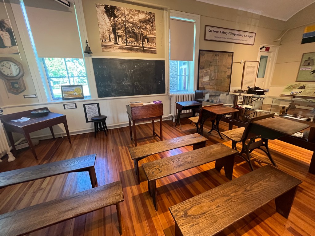 An exhibition staged as a classroom with wooden desks and benches facing a black chalkboard. 