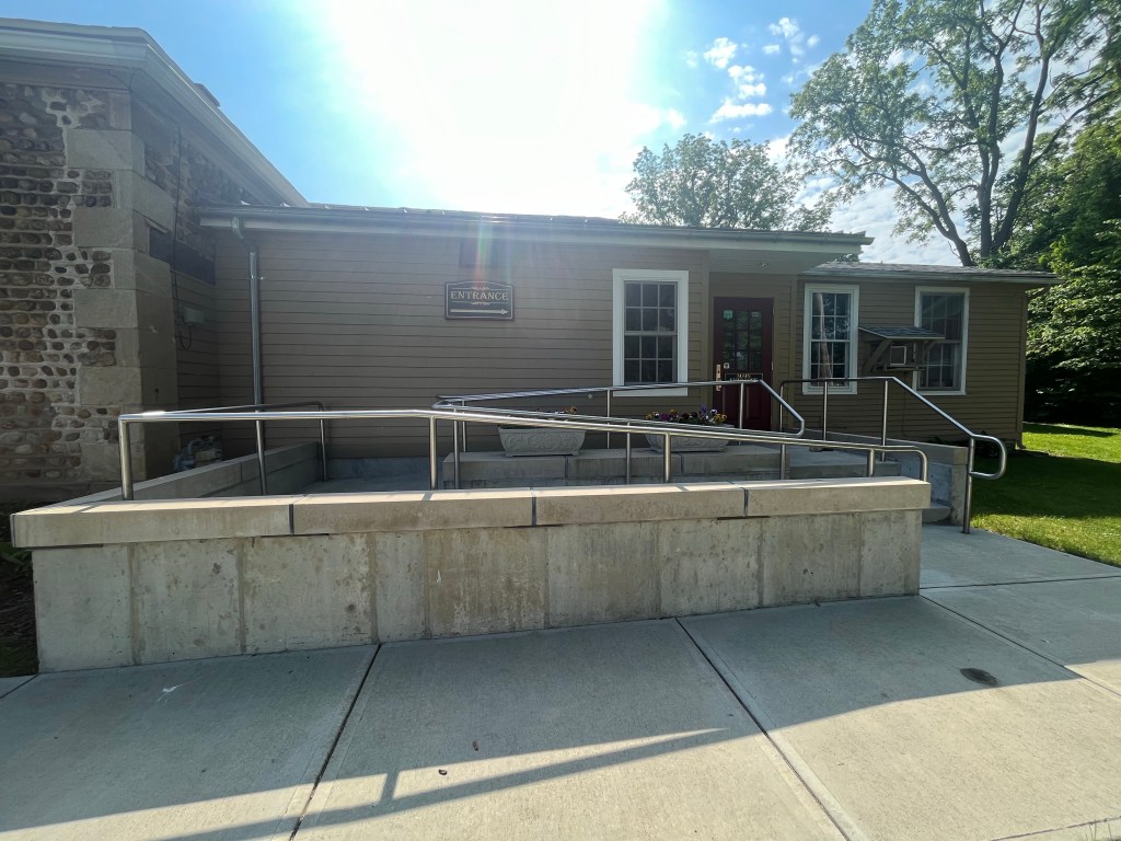 Main, accessible entrance to the Livingston County Historical Society & Museum, with concrete wheelchair ramp and stairway. An entrance sign points the way to a red door.